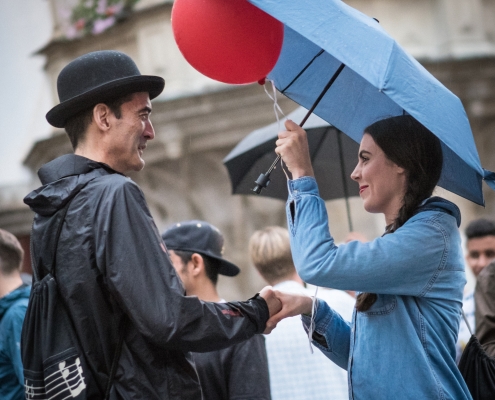 Ein Mann mit Hut gibt einer Frau, die einen roten Luftballon und einen blauen Regenschirm in der Hand hält, die Hand.